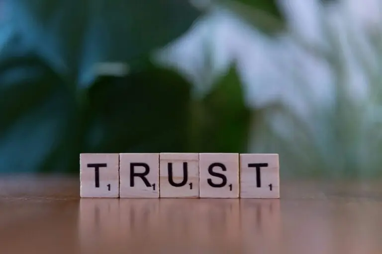 Four wooden Scrabble tiles spell out the word "TRUST" on a wooden surface, symbolizing how trust is essential in relationships and personal growth, with a blurred green background.