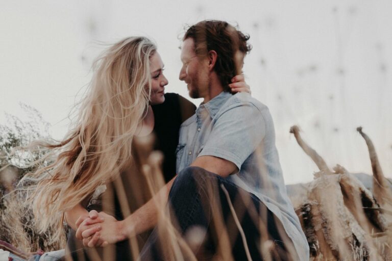 A couple sits closely together outdoors amid tall dry grass, gazing into each other's eyes and smiling, their bond reflecting the personal growth that enriches relationships. The woman's long blonde hair blows in the wind, with a soft, natural background.
