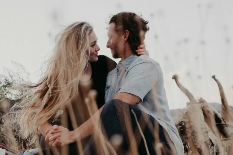 A couple sits closely together outdoors amid tall dry grass, gazing into each other's eyes and smiling, their bond reflecting the personal growth that enriches relationships. The woman's long blonde hair blows in the wind, with a soft, natural background.
