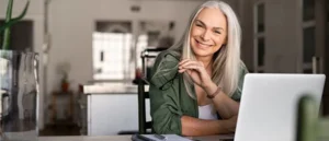 A smiling older woman with long gray hair sits at her bright modern table with a laptop and notebook, holding glasses, ready for a Small Group Live Coaching (Single Session) focused on self-improvement.