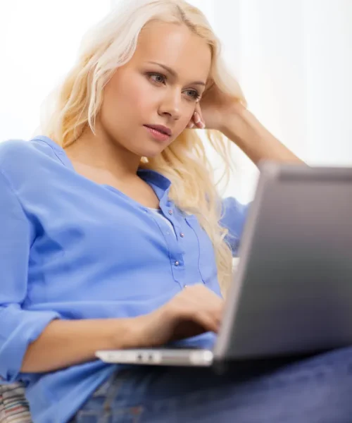 A woman with long blonde hair, wearing a blue shirt and jeans, is sitting and using a laptop, looking thoughtfully at the screen as she searches for coaching and support to strengthen her relationships.