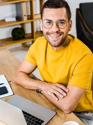 Smiling man with glasses and a yellow shirt sits at a desk with a laptop, smartphone, notebook, and coffee, looking up in a bright, modern workspace—a perfect setting for self-improvement and well-being.